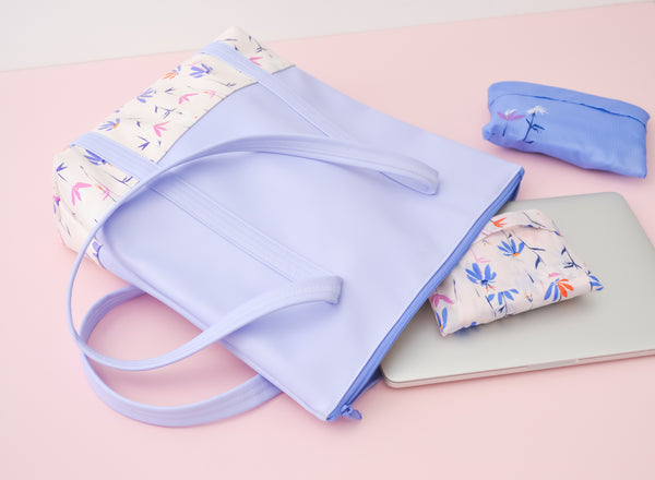 A periwinkle tote bag with magic sprigs on the bottom next to a macbook, and two packing cubes
