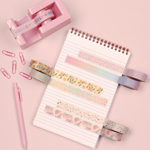 A notebook on a pink table with a pink tape dispenser, pens and paper clips. On the notebook six different washi take designs are taped onto the notebook page.