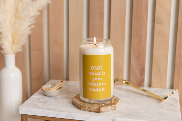 Can glass candle with yellow sticker that reads "OMG, what is your skincare routine" in pink lettering. Candle is sitting on a marble table surrounded by wooden matches and candle wick trimmer.