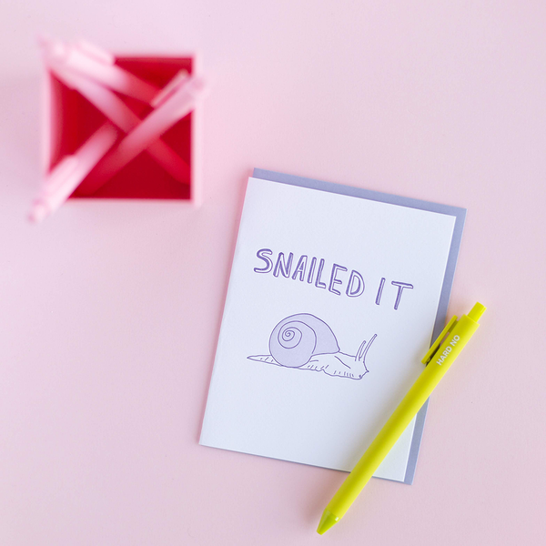 White greeting card with a purple snail and purple text "Snailed It'. There is a citron jotter pen with the text "Hard No" and a blush pink pen cup containing matching pens. The background is light pink. 