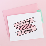 White greeting card with a pink and black banner and scripted writing "so many feelings". There is a pink envelope and a pink background.