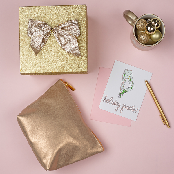 a 'holiday pants!' greeting card surrounded by a golden gift box, golden medium bag, golden mug and golden pen.