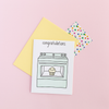 White greeting card with a mint oven. The oven has a clear window with a smiling muffin in it. The text at the top is 