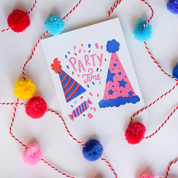 A white greeting card with a red and blue striped party hat, a pink with blue stars party hat and a pink and white wrapped piece of candy. There is red, blue and pink confetti with the text "Party Time".