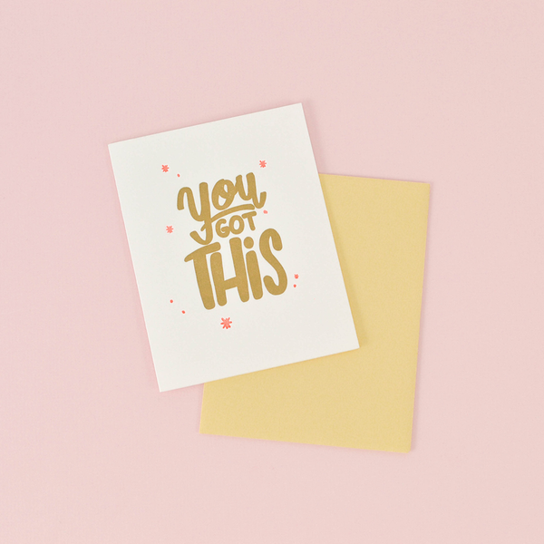 White greeting card with gold text "You Got This" and pink stars. There is a yellow card and a pink background.
