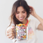 Cute, smiling young woman holding out a funny coffee mug that says So Many Feelings and is filled with colorful cereal.