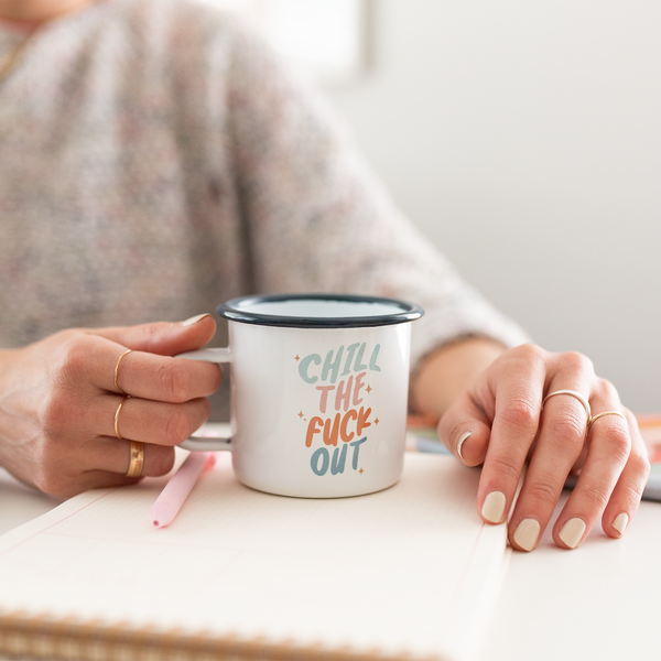 woman holding a A ceramic white mug with the phrase "Chill the fuck out" on it in pastel coloring. Rim of mug is a navy blue.