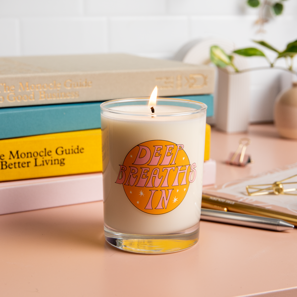Rocks glass candle with orange illustration and text that reads "DEEP BREATHS IN" in a pink font; candle is sitting on a desk surrounded by books and other desk items.