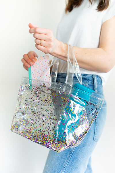 A woman holding a clear vinyl tote bag with multi colored confetti inside pulling out a mini notebook with a blue steel tumbler in her bag