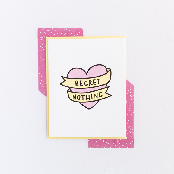 White greeting card with a pink heart and yellow banner, both outlined in black. The banner has the text "Regret Nothing". There is a yellow envelope and two small pink and white gift bags in the backround.