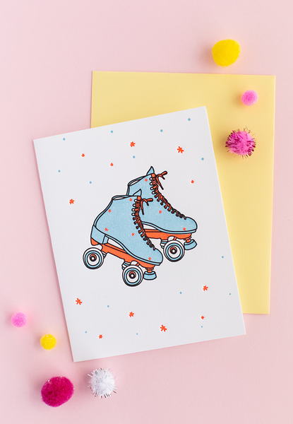 White greeting card with blue and coral/orange skates with dots and stars. There is a yellow envelope and colorful poms in the background.