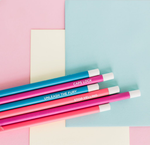 Six pencils in bright blue, pink and neon coral with different phrases printed and laying on colored paper.