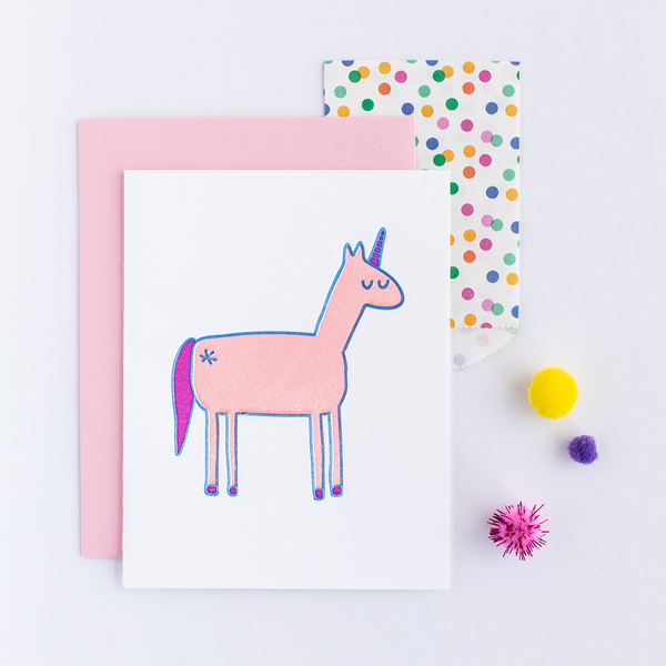 White greeting card with a light pink, dark pink and blue unicorn. There is a light pink envelope, a multi colored polka dot small gift bag and colorful poms. 