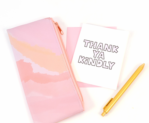 A white greeting card with light pink and black outlined text "Thank Ya Kindly" with a heart over the dot in the "i". There is a pink envelope, a gold jotter pen and a pink watercolor vegan leather zippered pouch.