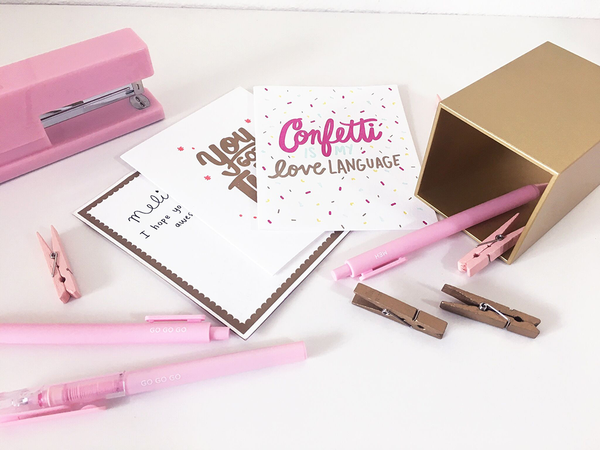 A collection of white greeting cards surrounded by pink and gold desk set items. The card on top has the text "Confetti is my love language" in pink, light blue and gold. The text is surrounded by multi colored confetti. 