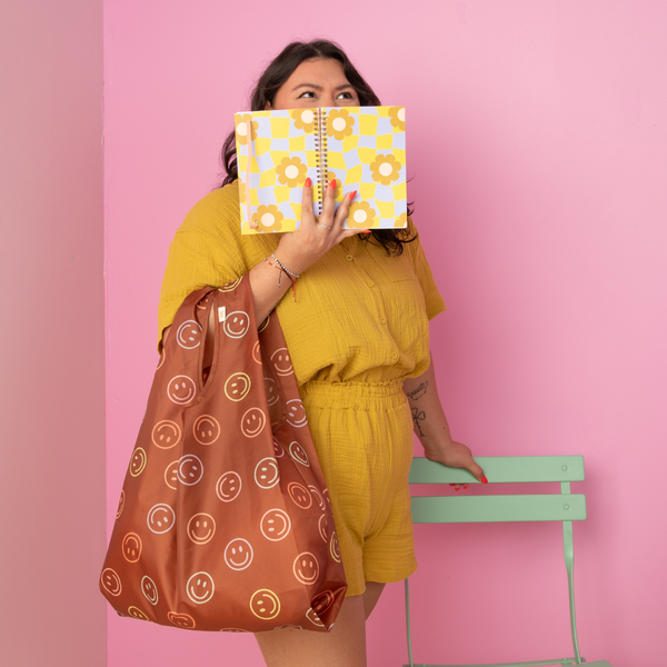 Pink background with a girl holding the cool funky daisy notebook and a brown smiley tote.
