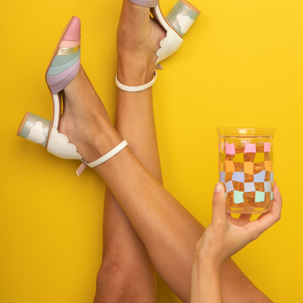 Carnival Checker glass tumbler being help up by a girl wearing rainbow and cloud heels with a yellow background.