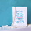 A white greeting card with a blue and pink clam opened to show a pearl. The blue script text above the clam is 