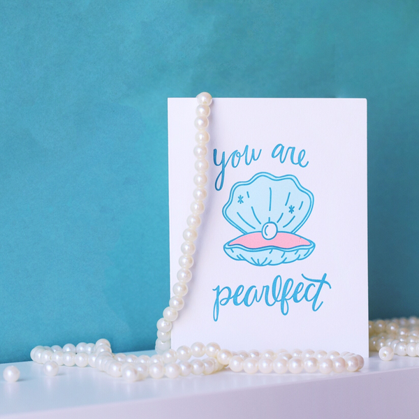 A white greeting card with a blue and pink clam opened to show a pearl. The blue script text above the clam is "you are" and below the clam is "pearlfect". The card is standing open with pearls hanging on it and a blue wall in the background.