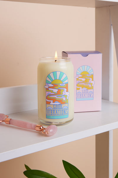 16 oz candle with "Good Vibrations" on it and a melting sunset. It is stood on a shelf next to a rose quartz face roller.