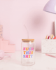 A clear glass tumbler with a bamboo lid and a curved glass straw sits on a white desk with a pastel pink background. Bold, colorful text on the glass reads “FUCK THIS SHIT” in pink, orange, blue, and purple letters. The aesthetic is playful and modern, with soft pink office accessories and a lavender desk lamp in the background.