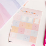 Sheet of planner tabs in "Sunset Stripes" color way, text with months of the year on each tab in white sitting on top of "Sunset Stripes" planner and surrounded by various stationery items.