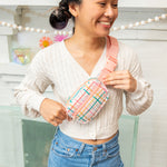 A girl wearing a pink colorful plaid small hip bag around her body with misc items in the back.