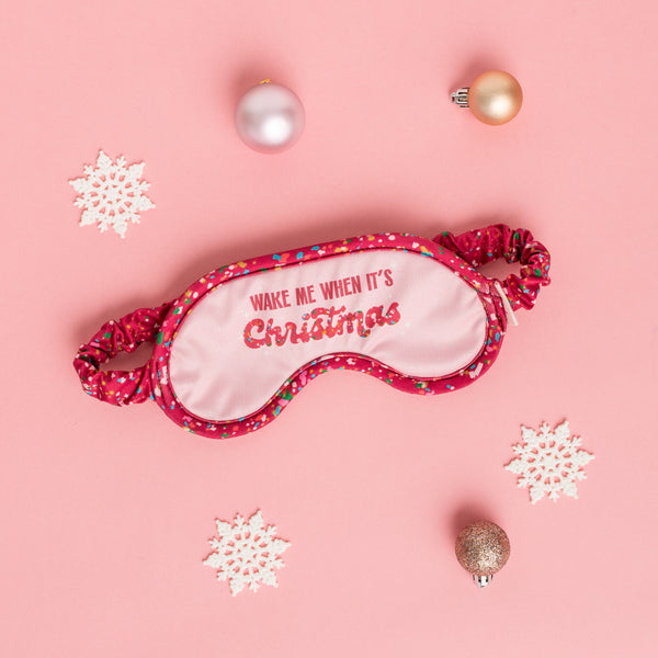 sleep mask with the phrase wake me when it's christmas with pink background on the boarder and band is red with mulit-color confetti detail.