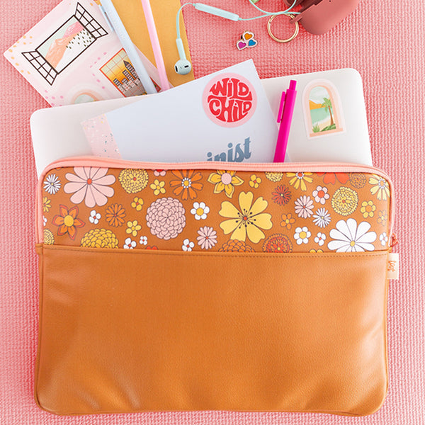Flower Power laptop sleeve with 1/3 retro flower pattern, and 2/3 orange-brown color. There is a notebook, laptop, pens, sticker sheet, and wired earphones coming out of the laptop sleeve, and it is against a pink cloth background.