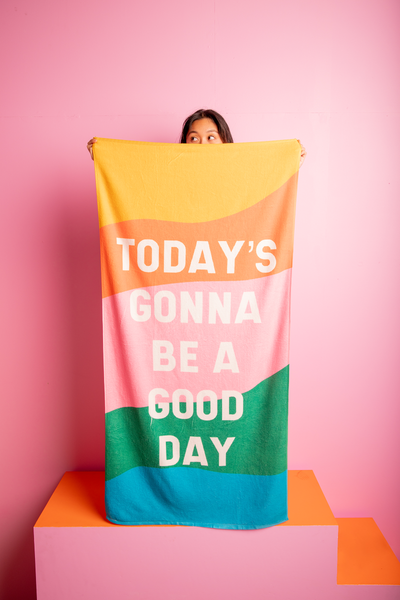 Girl on a pink/orange stair holding an all caps "today's gonna be a good day" text with colorful wavy rainbow beach towel. Pink background.