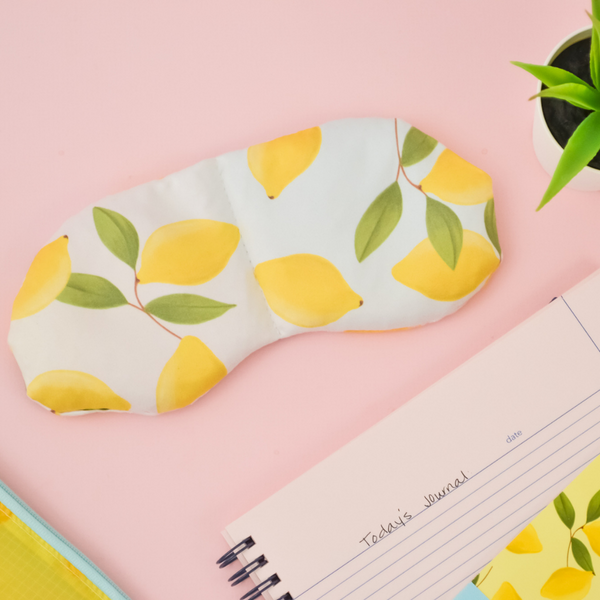 A weighted eye mask with lemons on it and a white and blue ombre background, surrounded by a journal with "Today's Journal", a succulent, a lemon ripstop pouch, and lemon mini notebook. 