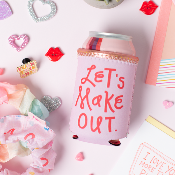 Let's Make Out Reversible Can Cooler is pink with Let's Make Out and a red side that says Don't Talk to Me in pink. Displayed with scrunchies, a small plant, stickers, and Jotter pens.