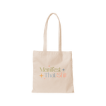 A cream-colored canvas tote bag that says, "Manifest that shit" on the front in different colored lettering. Phrase is surround by minimalistic sparkle stars. 
