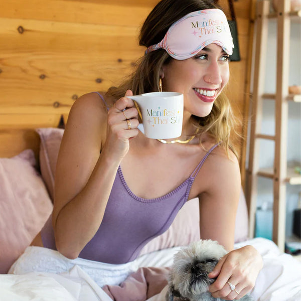 Woman playing with her puppy wearing Multi-Colored Sleep masked with band that reads Manifest That Shit. Includes an abstract squiggle pattern on the trim and head band support atop of her head.