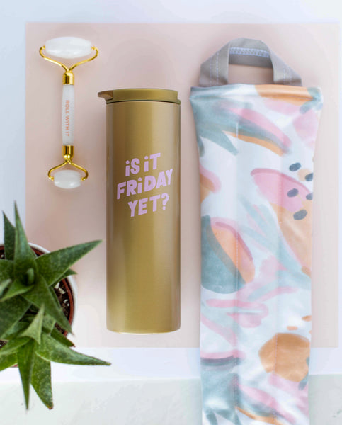  A weighted neck wrap with mutey fruity print laying next to a gold steel tumbler that says is it friday yet, and a white stone roller