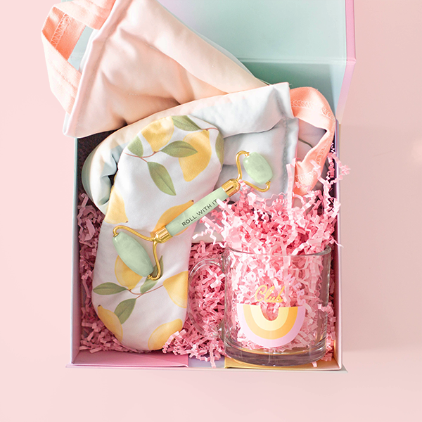 A gift set containing a clear glass mug with Daydreamers Club written in blue, a neck wrap with a soft rainbow gradient print, a lemon printed eye mask, and a jade face roller all in pink crinkle paper and packaged in a colorful box.