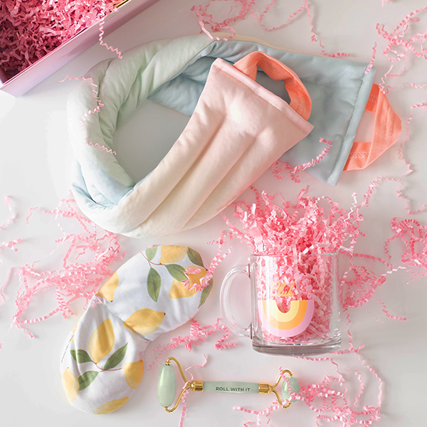 Contents of a gift set laid out to show a neck wrap with a pastel ombre print, a weighted eye mask with lemons printed on it, and a jade face roller amidst pink paper shreds.