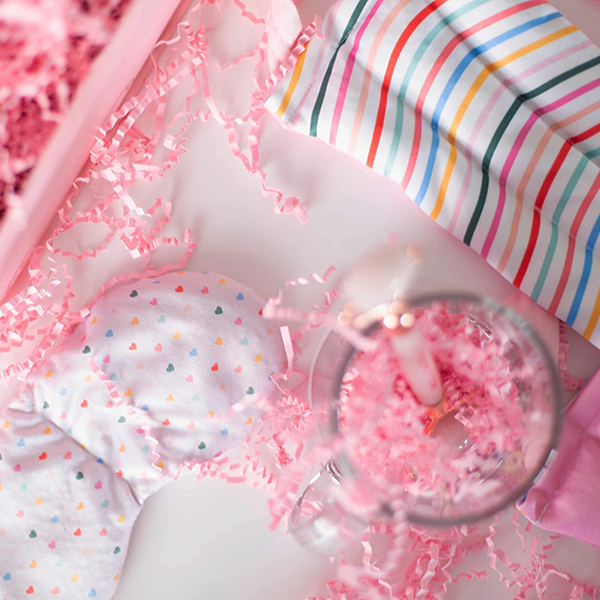 Contents of a gift set laid out to show a neck wrap with wavy rainbow lines, a weighted eye mask with tiny hearts printed on it, and a rose quarts face roller in a clear glass mug amidst pink paper shreds.