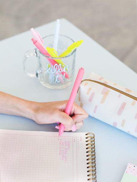 A person writing with a neon coral Jotter pen on a task pad. A glass mug with phrase "Kilin it!" is also displayed on surface with Citron green, blush pink, neon coral, and powder blue Jotter pens inside the mug.