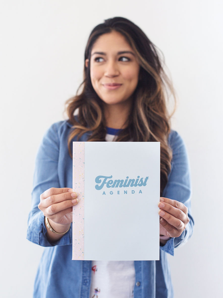 Cute young woman holding a small light blue notebook in front of her that says Feminist Agenda.