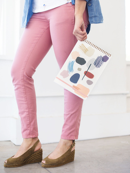 A woman in pink pants holding a large taskpad in one hand with an abstract art print on the cover.