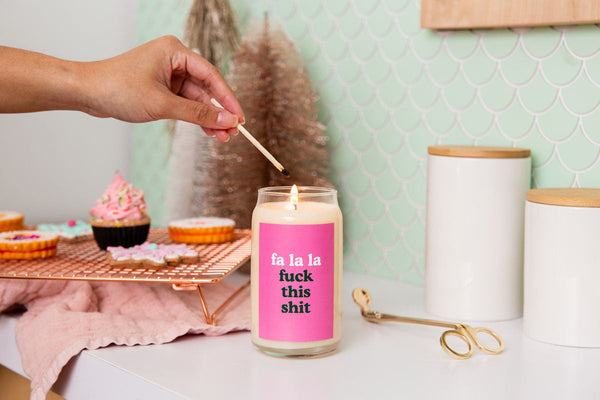 A 12 oz. holiday candle with a pink decal that says "fa la la fuck this shit." Candle is being lit with a match.