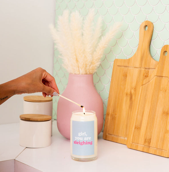 A 12 oz., "girl, you are sleighing" candle displayed on a white surface with someone lighting the candle with a match.