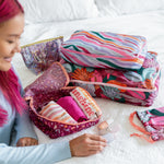 A small, medium, and large packing cube set which folds into a pouch. One with cranberry speckle, one jewel tone stripes, and one floral collage. Small cube is packed with multiple items and all cubers are displayed onto a bed with a confetti sparkled pouch next the the packing cube set.